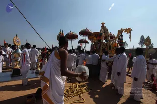 Outdoor temple procession on bare ground under a clear blue sky. A man in a white dhoti with a gold-bordered shawl holds a round silver drum-like vessel in the foreground. Behind him, a richly decorated chariot with floral garlands, tassels and a gilded ornamental canopy is surrounded by men in white shirts and dhotis. Several ornate umbrellas in red and multicoloured patterns rise above the procession, and a ceremonial brass vessel with a gold stand and red ribbons stands on a metal table to the left. A rope lies coiled on the ground and a small flame glows near the chariot. No clearly identifiable a person is visible.