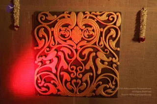 A decorative carved wooden panel with gold-painted scrolling foliate and floral motifs is displayed on a burlap-textured backdrop. Red light falls from the left, catching the raised gold scrollwork. A strand of white jasmine garland with red flowers hangs at each upper corner of the panel. No people are visible. The image is a photograph of a decorative art object.