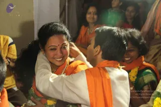 A candid photo at an indoor evening gathering: a smiling woman with hair pulled back and a red bindi wears a white top with an orange shawl and a gold necklace, embracing a young man in a white kurta with an orange shawl. Both have garlands of yellow and white flowers at their necks. Behind them, several smiling women in orange and green saris and a man in yellow watch.