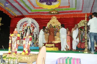 Outdoor evening ceremony on a decorated stage under a striped canopy with red, green and orange fringe. A seated man in orange robes and a rudraksha-style bead necklace sits on an ornate gold throne, with garlands draped on its sides. Two men in white dhotis stand near him, one leaning in beside a man in a white shirt; a bald man in white and an orange-clad man stand to the right. A cameraman with a tripod films at right. Behind the throne is a deity murti with multiple arms, a silver backdrop, and nearby female figures; in front, two decorated murtis with flower garlands rest on a gold tray with brass pots and oil lamps. A striped cloth lies on a table at lower right.
