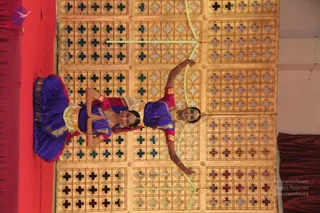 Two female dancers in blue and magenta traditional costumes perform on a red-curtained stage in front of a gold lattice backdrop with cross-shaped cutouts. The dancer on the left, in a deep blue skirt with gold border and gold waist belt, kneels with arms extended; the dancer on the right, in magenta and blue with gold jewellery and a headpiece, holds a long green garland drawn like a bow and arrow.