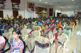 A wide view of a large hall filled with rows of seated women in colourful saris on white plastic chairs, facing toward the front. A golden ornate throne and a golden canopy shrine stand near the stage area, with framed portraits and photographs on the pale walls. Several women in the foreground have hands pressed together in prayer. Ceiling lights and air conditioners are visible. The setting suggests a satsang or discourse gathering.