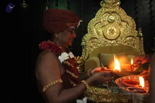 A person in a rust-red turban and bare-chested red-brown garment, wearing a red and white flower garland, gold armlets and bangles, holds a lit oil lamp on a brass tray in front of an ornate gold throne with a chair-like seat and carved backrest. Another hand holds a lit lamp at the right edge, and a woven basket holding a lit diya sits at lower right. Dark background with hanging brass bells. Swamiji (probable): the figure wears a red turban, rudraksha-style garland and gold ornaments, presiding over the lamp offering as a priest-like figure.