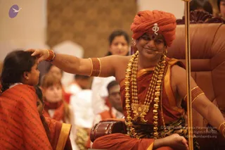A photo of a seated figure on an ornate brown leather throne, wearing an orange-red turban with a silver emblem, an orange-red robe with gold border, a multi-strand gold necklace with a large gold pendant, gold bangles and a rudraksha-style mala, and a lapel microphone. Their right arm extends to rest a hand on the head of a young girl in a red-orange shawl with a gold border, who sits close in front. Blurred seated devotees in red and white clothing fill the background. Probable Swamiji cues: turban and saffron/orange robe with ornate gold necklaces, seated on a throne during a satsang-style gathering.