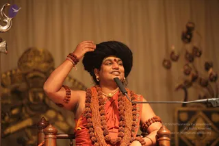 A seated man in an orange-saffron robe and a long orange-and-rudraksha mala stands at a wooden chair before a microphone, raising one arm to his head with a smile. He wears a black swept-back hair style, gold earrings, a gold necklace, rudraksha bracelets, and a yellow tilak with red dot. Behind him, a large golden deity murti and a dim curtain with decorative metal lamps form the stage backdrop.