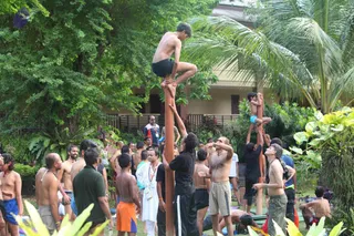 Outdoor photo at a tropical garden retreat with palm trees and a low building behind. Several shirtless young men and boys climb tall wooden poles, one boy in black shorts at the top balancing with his feet on the pole. Around them a crowd of men in shorts, tank tops and a white dhoti-style garment stands watching, with one man in a green shirt and another in a white kurta with a lanyard. A man with grey hair and a white beard stands on the right in shorts and a grey shirt, arms raised toward the pole. The scene appears to be a physical training activity among participants. The ritual or yoga element is not visibly confirmed beyond the poles and crowd.