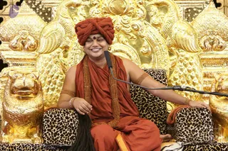 A smiling man in saffron-orange robes and a matching red-orange turban sits on an ornate golden throne carved with lions and floral motifs, with leopard-print cushions. A microphone on a stand is positioned near him, and he rests one arm on the armrest. He wears a rudraksha mala draped over his shoulders, bracelets on his wrists and upper arms, and a red-orange wrap. Swamiji (probable): long hair and ornate saffron attire with a turban and rudraksha mala on a throne, consistent with a satsang discourse setting.