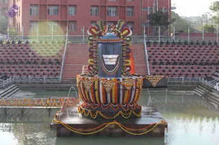 A large Shiva linga stands on a square platform in the middle of a water pool, decorated with dense garlands of marigold and multicoloured flowers in red, yellow, purple and white. A wide band of garlands wraps the base, and orange-and-yellow flower strings hang along the platform edge and a footbridge. Water jets spray around the platform. Behind the linga, a stepped seating area with rows of empty chairs leads to a red multi-storey building. A few small figures stand on the raised steps above the linga, too distant to describe in detail. The scene suggests a Mahashivaratri abhishekam-style display of a decorated deity murti.
