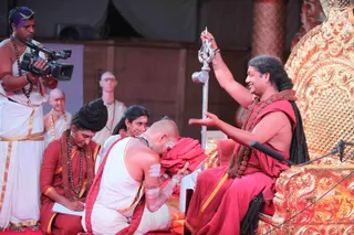 A photograph of a ceremonial initiation on a stage. A seated man in red robes with long swept-back hair and a dark rudraksha mala raises a silver trident-like staff with his right hand and gestures with his left, sitting on an ornate gilded throne with carved golden panels. A shaven-headed man in white clothes with orange borders and white body markings kneels before him, bowing with his head near a folded red cloth. Behind them, a woman in white with a red border sits beside a man in red with a mala who writes on a clipboard, while a cameraman in white with a camera on his shoulder films, and two shaven-headed men in white stand in the background. Five people are visible.