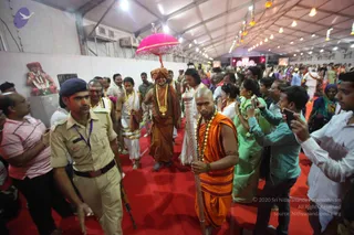 A procession walks down a red carpet inside a large white marquee lit by strings of hanging lights. In the centre, a man in an orange robe with a bare shaved head and a long garland of flowers carries a tall silver staff. Behind him, a figure in a saffron-brown robe with a large gold necklace and a tall orange turban walks beneath a pink fringed parasol. Men in white dhotis with gold-bordered shawls and garlands walk nearby. A uniformed police officer in khaki with a rifle stands at the left edge, and a dense crowd of devotees in saris and kurtas, many holding up phones to film, lines the carpet. A deity figure decorated with garlands sits on a raised shelf at the upper left.