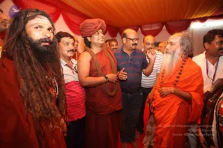 Photograph of a crowded tented gathering under a red and orange canopy with draped red fabric. In the centre, a man in a rust-red robe wears a rust-red turban and a thick brown bead necklace, smiling as he listens. To his right, an older bearded man with long white hair and an orange robe strung with rudraksha beads gestures in conversation. On the left, a man with long dreadlocks and a beard wears a rust-red wrap. Around them, men in striped polo shirts, a checked shirt and white clothes stand close together on a red carpet. The centre figure and the bearded man in orange are the only ones with distinctive ceremonial cues, so the identity of the centre figure is uncertain.