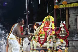 A bare-chested priest in white dhoti with an orange-saffron shawl stands at night before a decorated palanquin-like ratha or peetha heavily garlanded with yellow and red marigolds and white jasmine strands. A small oil lamp with a flame and a brass lamp stand flank the shrine, and the priest holds a lit lamp or aarti offering toward the decorated structure. A cameraman with a still camera and another with a video camera on a tripod film the ritual. Strings of lights and a yellow-fringed canopy hang overhead, with a carved stone pillar on the right. The scene suggests a puja or aarti with decorated vahana.