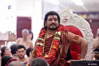 A man seated on an ornate silver-and-red throne at a satsang, wearing a rust-brown dhoti and shawl, multiple garlands of red and white flowers, gold beaded necklaces, gold armlets and a rudraksha-style ornament at his neck. He has a red bindi and a sideways gaze, with a red cushion behind him. Audience members in white and rust clothes sit in the foreground and background, and a YAMAHA-branded device is visible at lower right. a person appears as the seated figure on the throne, distinguished by the ceremonial garlands, gold ornaments and dark swept-back hair. Seated devotees surround the dais.