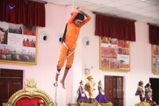 A young performer in an orange long-sleeved top and orange trousers climbs a white rope suspended from above, body raised off the floor with bare feet and legs wrapped around the rope, arms overhead gripping it, a white headband on the forehead and long dark braids flying. Behind, framed photographs of a river gathering in saffron-clad crowds hang on a pink wall beside red curtains. Below, a golden ornate throne and several decorative figures in purple and blue garments with a small golden deity figure stand on the stage.
