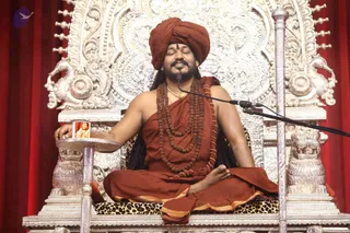 A seated man in an ornate silver throne with lion-head armrests and a leopard-print seat cushion, against red drapes. He wears a rust-orange turban, a rust-orange shawl and dhoti with gold border, multiple rudraksha malas, and a gold chain, eyes closed, smiling, legs in lotus posture, with a microphone on a stand before him. His left hand rests on a silver tray holding a small framed photo of a man with a long beard. Swamiji (probable): a long-haired figure in orange turban and rudraksha malas seated on the throne giving satsang, with distinctive ceremonial cues matching the page.