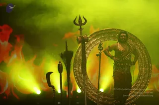 A staged Kalabhairava-style performance under green and orange stage lighting with flame backdrop. A single man stands at right with hands on hips, bare-chested, wearing a dark patterned dhoti, rudraksha-style beaded necklaces, arm bands and bold white facial markings, with a large golden ring behind him. Ceremonial props stand in front: a tall trident, a curved sickle-like blade, a ceremonial staff with a tufted fan and a small ornate sceptre. Smoke fills the stage. The dramatic pose and props suggest a deity-form presentation rather than a clearly identifiable a person; appearance cues are not distinctive enough to confirm identity.
