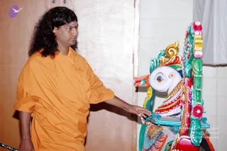 A photograph of a man with long, curly dark hair and a red bindi on his forehead, wearing a loose orange-saffron robe, standing at the left and touching a brightly painted wooden vehicle figure of a bird-headed mount with a white head, orange beak, and turquoise, red, yellow and green ornamental carving. The figure sits beside a white tiled wall and a white cloth hanging. Probable Swamiji: the long hair, orange robe and presence at a painted ceremonial vehicle match the page's Swamiji reference, though the face is not used for identification.