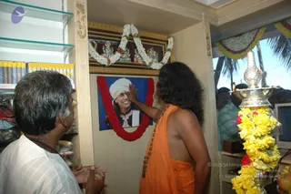 A photograph inside a shop interior where a bare-chested man with long, dark, swept-back hair and an orange saffron dhoti, wearing a rudraksha mala, reaches up to touch the forehead of a framed portrait. The portrait shows a smiling man in a white turban and ornate dark embroidered garment against a blue background, garlanded with a red and white flower garland. Above it hangs a gilded frame with a garland and silver crown-like ornaments. To the left, a man in a white checked shirt with a rudraksha mala watches with hands together. A tall silver lamp stand wrapped in yellow marigold garlands stands at right, and a second person is visible in a light green shirt behind it. Glass shelves hold stacked books and cloth goods.