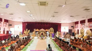 Wide view of a hall set for a satsang or puja. At the centre, a seated figure in saffron-orange robes sits on a golden throne under a gold canopy, flanked by golden elephant and lion figures and attendants. Standing attendants in white and one in purple move around the dais. Rows of seated devotees in saffron and orange robes, some with shaved heads, fill the floor on both sides, with a long green-and-white floral rangoli leading toward the throne. Red drapes and framed pictures line the walls, and a gold deity murti stands at the right.
