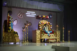 A stage set for a satsang, with a seated figure on an ornate gold throne with red cushions, wearing a blue garment and a white and orange flower garland. Only one person is visible on the central throne. Another golden throne stands empty on the left, draped in marigold and multicolour garlands and a blue-clad figure is depicted on its backrest. Brass lamps with lit flames stand on both sides, and a red and yellow canopy hangs above the central seat. The backdrop has a white Shiva lingam emblem, a trident motif and a dove symbol on draped purple fabric. A microphone stands beside the throne.