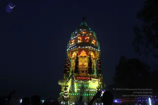 Night photograph of an illuminated festival ratha (chariot) lit with warm yellow, orange and green lights, its tiered, pyramidal superstructure topped by a kalasha-like finial. A deity murti with a crown is framed in the central niche, flanked by garlands hanging from the sides. A white-clad figure stands on the chariot platform near a fan-like ornament, and silhouetted devotees in the foreground hold up phones to record the procession. Strings of coloured lights decorate the ground at right under a dark sky.