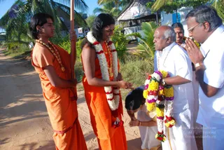 Outdoor photo on a sandy dirt path lined with palms and thatched huts. A young man in an orange robe and orange shirt, holding a tall wooden staff, stands at left with a gold-bead necklace. Beside him a figure in a saffron-orange robe with a long swept-back hair, wearing a white and red flower garland, a gold ceremonial necklace and rudraksha beads, stands with a hand extended. A man in white dhoti and shirt with a multicoloured flower garland and a rudraksha mala bows forward, and a kneeling person in white touches the ground in obeisance. Another man in white with folded hands stands at right, and a smiling woman in white is partly visible behind.