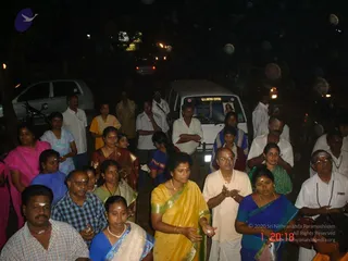 Night photograph of a crowd of devotees gathered on a street at a deity procession in Chennai, Tamil Nadu. Many people stand with palms pressed together in namaskar or holding folded hands. A woman in a yellow sari with gold border and a woman in a blue sari with a white shawl stand near the front, and a man in a white shirt and light trousers with a rudraksha mala and a second man in white with vibhuti on his forehead stand nearby. Parked vehicles, a white van and street lights are behind them, with bokeh light circles in the frame. Counting the visible people, there are more than twenty.