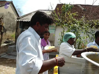 Outdoor relief food distribution in a village courtyard with tiled-roof houses and a climbing vine. A man in a white shirt with a red wrist thread holds a paper plate of food while a young woman in a purple top stands behind him. A man in a white shirt and green hairnet serves food from a large metal pot onto plates. Another server in white stands at right, and a steel tub of rice sits on a white table.