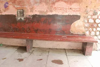 A weathered red-painted wooden bench stands against a peeling pink plastered wall with a rough stone wall section to the right. Above the bench, Devanagari script is painted on the plaster, and a small metal plaque is mounted on the wall at left. The stone floor shows dark stains. No people are visible; the scene is an empty courtyard-type corner, possibly near a temple or ghat area.