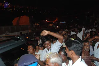 Night crowd scene at a roadside event, with many people pressed close around parked cars and a vehicle window. A smiling young man in a white shirt raises his arm high in the centre, while others in white clothes and saffron-toned garments reach up around him. Some people have flower petals and yellow blossoms on their heads and shoulders. Strings of small lights and bunting glow in the dark background, with a dim building wall on the left.