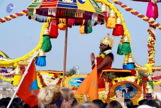 A festive procession on a decorated chariot (ratha) under a multicoloured canopy with tassels and gold bells, strung with yellow and pink flower garlands. A standing man in saffron-orange robes wears a gold crown and a white flower garland, his palms joined in greeting namaskar. He holds an orange flag post, and a crowd of devotees, mostly seen from behind, raises hands in the foreground. Appearance and role suggest the presiding figure of the procession, a person, but the face is not clearly visible. Orange and yellow flags and the chariot's floral decoration fill the scene.