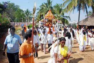 Outdoor procession on a dirt path lined with palm trees and a thatched hut. At the centre, a decorated ratha with a multicoloured canopy and a deity murti is pulled forward. Around it, devotees in white clothes with yellow and orange sashes walk, some holding a long brass staff and a trident. A man in an orange shirt with a lanyard leads on the left, while a woman in a yellow-bordered white sari and a man in a light blue shirt are visible in the foreground. A cameraperson films on the right.