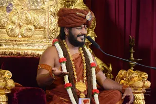 A smiling man in rust-orange robes sits on an ornate gold and red velvet throne, speaking into a microphone on a stand. He wears a rust-orange turban with a jewelled brooch, a dark rudraksha mala, a heavy gold ceremonial necklace, and white and red flower garlands. His right hand is open in a gesture while he talks. Behind him are gilded carved panels and a red curtain, with a lamp stand visible at right. Swamiji (probable): a bearded man in saffron-orange turban and robes, with rudraksha mala and gold necklace, seated on a golden throne during a discourse.