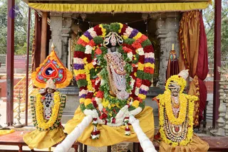 A decorated deity murti is seated at the centre of a shrine canopy, framed by a large arch of marigold, rose and green-leaf garlands in yellow, red, purple and white. The central figure wears ornate silver-and-gold jewellery and a tall crown-like headdress, seated on a yellow cloth. Flanking statues on each side wear yellow and orange garlands and ornate jewellery; the left figure is seated beside an ornamental orange-and-red umbrella-like canopy. Stone pillars, red curtains and a fenced area with a small sign appear behind. No living people are visible.