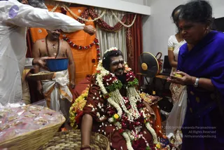 A seated man in a maroon robe is heavily garlanded with white jasmine strands and red and yellow flower garlands, with petals scattered over him, while smiling. A bare-chested priest in an orange-and-white dhoti with a rudraksha mala and gold chain stands behind him holding a blue bowl, and a man in white is pouring or offering from above. A woman in a blue sari with gold-bordered design holds a small yellow fruit, and another woman in white stands in the background. Behind them hang orange cloth and red-and-white draping with floral garlands, and a basket of packaged offerings sits in the foreground.