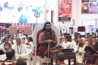 A man seated on an ornate silver-and-red throne at the centre of a hall, holding a silver trident-topped staff upright beside him. Around him sit many devotees in white and cream clothing and orange sashes, with dark blindfolds or eye coverings, many wearing rudraksha malas. A small wooden table in front holds a red tray, a microphone and an electronic keyboard. A screen behind shows colourful blue and pink imagery, and framed photos hang on the wall. Partially visible people sit in the foreground.