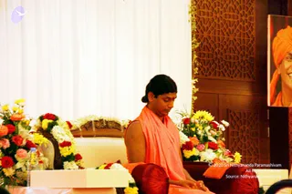 A man in a saffron-orange sleeveless robe and drape sits cross-legged on a cream upholstered seat with a carved wooden armrest, eyes lowered and hands resting in his lap. He has a red bindi on his forehead and dark hair tied back. Behind him a portrait of a smiling man in an orange turban hangs on a dark wooden panel, and red, yellow and white flower garlands decorate the seat and a side table holding a water bottle. A white curtain and a geometric wooden screen form the backdrop. a person is seated as the speaker of a satsang-style session, with a bare-shouldered orange robe and a red bindi as the appearance cues.