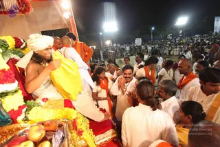 Night outdoor event with bright stage lights and a large crowd behind barricades. In the foreground, a figure with a white turban and bare upper body, wearing gold necklaces and a rudraksha-style adornment, sits on a red-draped throne decorated with yellow, white and red flower garlands; he holds a yellow cloth and smiles. Around the throne, devotees in white and saffron/orange shawls press forward, one man in white with folded hands smiling, a woman in white with an orange shawl, and fruit offerings sit on a gold tray at the bottom left. a person cues: turban, bare-chested with gold necklaces, seated on a throne being honoured by devotees.
