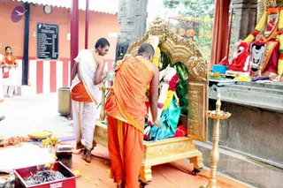 A temple ritual at a shrine with a gilded arched canopy and golden base holding a deity murti draped in green silk and garlanded with yellow and red flowers. A shaved-headed priest in an orange dhoti and orange shawl leans forward to adorn or attend the deity, while a second priest in white dhoti with a gold border stands beside him holding a lamp. Brass oil lamps stand nearby, with fruit, flowers and a red-cased fire pit with burning coals in the foreground. Behind them, a black board lists temple information in white text, and a red-and-white painted wall frames the courtyard.