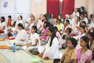 A seated group meditation session in a bright hall with pale walls, red curtains and a white door. Many participants sit cross-legged on thin mats with eyes closed, including young women in white kurtas and white dhotis, girls in pink and patterned salwar, and a woman in an orange shawl. Water bottles, plastic tiffin-style containers and a small brass lamp sit on the floor, and backpacks are stacked along the back wall.