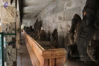 A photograph of a long, narrow stone colonnade inside a temple, with a raised stone plinth running along the right side holding a row of dark stone sculptures, including small deity figures and a seated figure. On the right, a large dark carved stone figure with a crown-like headdress and a red tilak mark on the forehead stands beside the plinth. The walls are grey granite blocks, the floor is smooth stone, and the pillars on the left are carved with reliefs and fitted with green metal grilles. No people are visible.