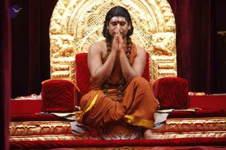 A seated man in saffron-orange robes with a yellow sash sits cross-legged on a tiger-skin-covered platform before an ornate gold-leaf throne back carved with lions. He has long swept-back hair, a white forehead marking with a red dot, and a long rudraksha mala, with his palms pressed together in namaskar and eyes closed in meditation. Red velvet cushions with tiger-stripe pattern flank him, with fruit offerings visible at the sides. a person, seated in a satsang setting. A red curtain frames the scene.