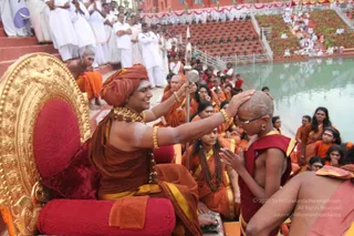 Outdoor ceremony beside a stone-edged water tank at a temple complex. A seated figure in a red-orange turban and ochre robe, with ornate gold necklaces and bangles and white markings on the forehead and arms, rests a hand on the bald head of a kneeling, bare-chested young disciple in maroon and gold cloth. The seated figure holds a long ceremonial staff with a silver top. Behind them stand saffron-robed devotees, some with rudraksha malas, and a crowd in white on stone steps and terraced seating under red and white bunting. A gilded throne with red cushions occupies the left foreground.