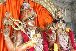 Two metal murti statues of deities stand before an orange silk backdrop with a gold arched frame. The figure on the left, in a striped red and gold turban with beaded tassels, holds a trident and a snake-topped staff in one hand and a gold mudra hand raised; he wears rose garlands, rudraksha-style beads and a white beaded necklace. The figure on the right wears an ornate jewelled crown, large earrings and layered pearl and stone necklaces, and holds a small flame-like object and a ritual item. Both are adorned with red rose garlands and yellow marigolds. No physical people are visible.