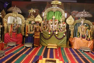 A photograph of a temple-style shrine with several gold-framed thrones and canopies draped in green, red and orange cloth. Multiple deity murtis adorned with gold necklaces and garlands of white and orange flowers sit on the thrones. A standing man in an orange-brown turban and saffron-brown dhoti-style garment with a gold-and-white garland stands centrally before the shrine, one hand raised. Striped woven mats cover the floor, with brass boxes and a low metal stand in front. The backdrop shows painted temple gopurams.