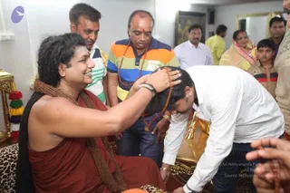A seated man with long, swept-back dark hair and a bare-shouldered rust-red robe, wearing heavy brown rudraksha mala necklaces, places his right hand on the head of a man in a white shirt who bends forward. a person: the seated figure has long swept-back hair, a rudraksha mala and a rust-red robe, consistent with the page's subject. Behind them, several onlookers stand in a bright room: a man in a striped polo with a rudraksha mala, a man in a striped shirt, a man in white, a woman in a yellow and pink sari, and a boy in a striped shirt. A gilded chair with a leopard-print cushion and a marigold-and-tassel garland sit at the left edge.