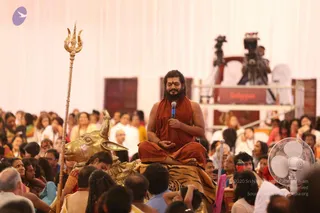 A seated man in an orange-red robe sits cross-legged on a golden throne draped with a tiger-striped cloth, eyes closed, holding a handheld microphone in his right hand. Beside him stands a large gold bull figure and a tall golden trident. Around him a large crowd sits on the floor in saris and white clothes, with a camera on a tripod and a white fan in the foreground. A white draped stage backdrop and a red-and-gold structure appear behind.