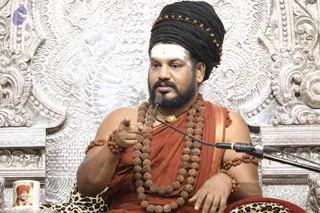 A seated man in a dark turban wound with rudraksha beads and a white vertical tilak on his forehead speaks into a microphone while pointing his right hand forward. He wears a rust-orange shawl over bare shoulders, a long multi-strand rudraksha mala, and a gold pendant, with rudraksha bracelets on both wrists. He sits on a leopard-print cushion before an ornate silver embossed throne backdrop. In the lower left, a white mug printed with a turbaned man's portrait and the appears. The setting suggests a public discourse or satsang