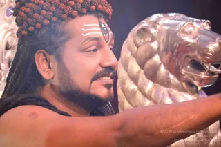 A bare-shouldered man with long matted dreadlocks wrapped under a dense crown of rudraksha beads leans toward a large polished silver lion-head sculpture, his hand resting on it. He wears a white tripundra and a painted third-eye motif on his forehead, and a dark cloth drapes his shoulders. Warm stage lighting falls across his face against a dark backdrop. The pictured person has distinctive rudraksha and dreadlock cues, so this is a person appearance, shown in person as a sannyasi-style presenter beside the silver lion sculpture.