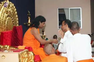 Indoor satsang scene in front of a golden ornate throne with red velvet cushions. A man with long swept-back dark hair and a saffron-orange robe sits on the throne, extending his hand toward a devotee. Two shaven-headed men in white clothing and an orange-robed attendant kneel before him. A brass oil lamp with a lit flame stands beside the throne and another lamp burns on a table. A microphone rests on a book. Behind them a person sits near a window with white curtains, and a wooden box with gold trim sits at the lower left. The appearance-based identity of the seated man is probable, with cues of long hair, saffron robe and a throne setting, so he is a probable Swamiji seated in person on the peetha. Four physical people are visible.